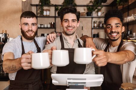 Group of cheerful men baristas wearing aprons working at the counter in cafe indoors, toasting with...