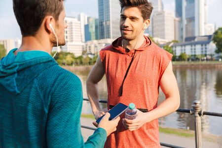 Photo of two strong sporty men wearing sportswear using cellphone and earphones while talking on...