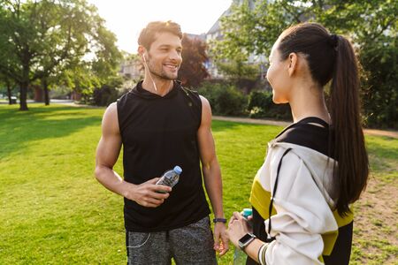 Photo of young nice man and woman wearing sportswear talking together and holding plastic bottle while working out in city parkの写真素材