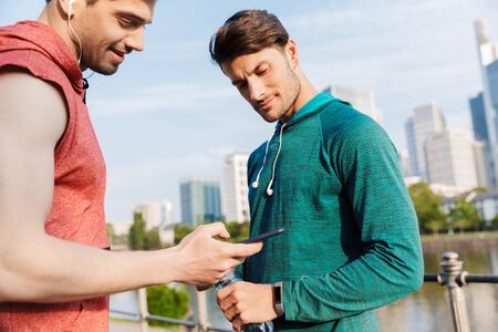 Photo of two young sporty men wearing sportswear using cellphone and earphones while working out on...