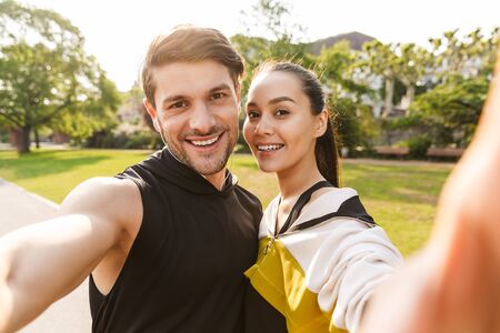 Photo of smiling pleased man and woman wearing sportswear taking selfie photo while working out in city parkの写真素材