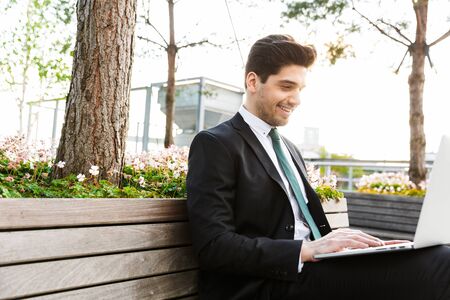 Attractive confident young businessman wearing a suit sitting on a bench outdoors at the city street, working on laptop computerの写真素材