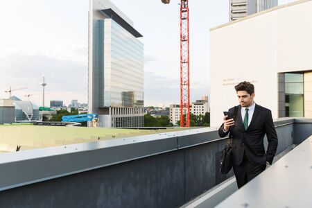 Handsome young businessman wearing suit walking outdoors at the city streets, using mobile phone