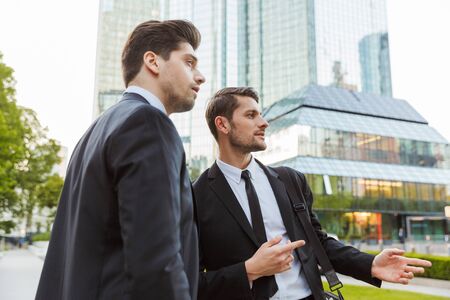 Two attractive confident young businessmen wearing suits standing outdoors at the city streets, discussing new projectの写真素材