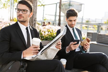 Two attractive smiling young businessmen wearing suits having a lunch break while sitting outdoors at the city streets, drinking coffee and reading newspaperの写真素材