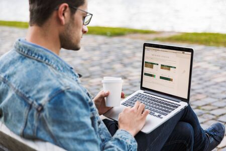 Side view of a handsome young man dressed casually spending time outdoors at the city, using laptop computer while sitting on a bench and drinking takeaway coffeeの写真素材