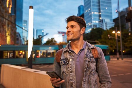 Attractive smiling young man walking at the city streets in the evening, using mobile phoneの写真素材