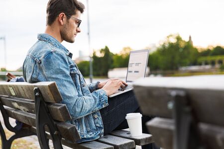 Side view of a handsome young man dressed casually spending time outdoors at the city, using laptop computer while sitting on a benchの写真素材