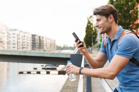Handsome smiling young man dressed casually spending time outdoors at the city, carrying backpack, using mobile phone while leaning on railの写真素材