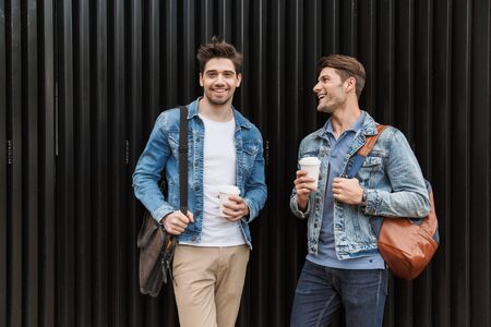 Two smiling young men friends dressed casually spending time together at the city, drinking takeaway...