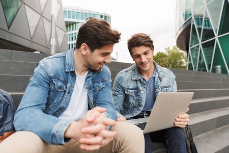 Two smiling young men friends dressed casually spending time together at the city, using laptop computer while sitting on staircaseの写真素材