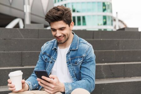 Handsome smiling young man dressed casually spending time outdoors at the city, sitting on stairs, holding takeaway coffee cupの写真素材