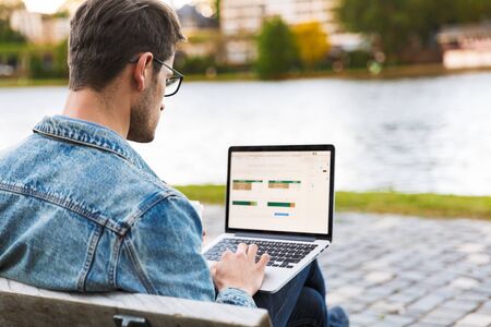 Side view of a handsome young man dressed casually spending time outdoors at the city, using laptop computer while sitting on a benchの写真素材