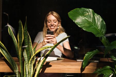 Portrait of blonde young woman smiling and holding smartphone while sitting in cafe with green plantsの写真素材