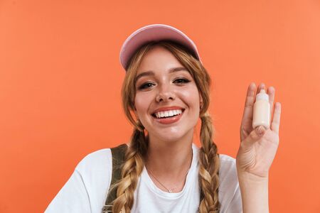 Image of joyful blogger girl wearing hat holding cosmetic bottle and demonstrating new product isolated over orange wall in studioの写真素材