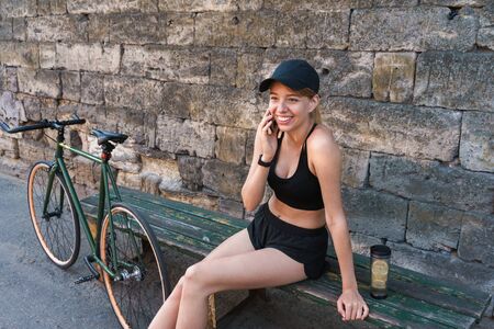 Image of cheerful blonde woman dressed in sportswear talking on smartphone while sitting on bench...
