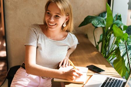 Portrait of optimistic young woman using laptop computer and writing notes in diary book while sitting in cafeの写真素材