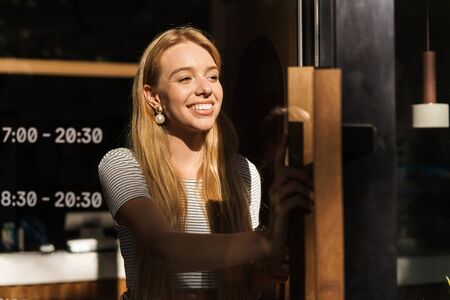Portrait of cheerful young woman smiling and opening glass door while leaving coffeehouse or cafeの写真素材