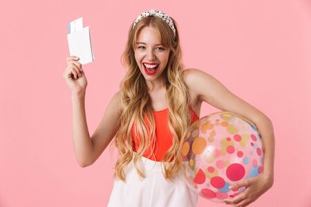 Image of happy young woman with long curly hair smiling while holding summer beach ball and travel tickets isolated over pink backgroundの写真素材