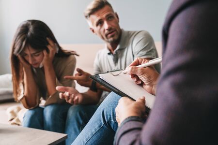 Photo of upset irritated couple man and woman having conversation with psychologist on therapy session in roomの写真素材