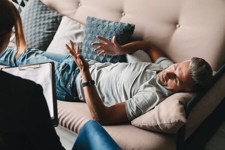 Photo of unshaven casual man lying on sofa and having conversation with psychologist on therapy session in roomの写真素材