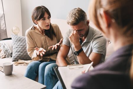 Photo of puzzled annoyed couple man and woman having conversation with psychologist on therapy session in roomの写真素材