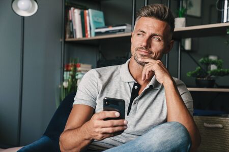 Photo of happy caucasian man in casual clothing holding smartphone while sitting on armchair at home