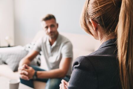 Photo of caucasian focused man having conversation with psychologist on therapy session in roomの写真素材