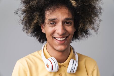 Photo of joyful caucasian man with afro hairstyle wearing headphones smiling at camera isolated over gray backgroundの写真素材