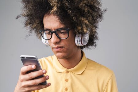 Photo of puzzled caucasian man with afro hairstyle wearing headphones and eyeglasses holding smartphone isolated over gray backgroundの写真素材