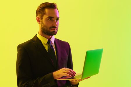 Picture of a concentrated young business man posing isolated over light green background wall with led neon lights using laptop computer.の写真素材