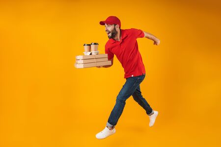 Portrait of young delivery man in red uniform running with pizza boxes and takeaway coffee isolated over yellow backgroundの写真素材