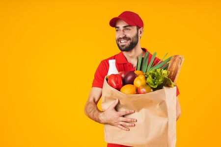 Portrait of attractive delivery man in red uniform smiling while carrying paper bag with food products isolated over yellow backgroundの写真素材