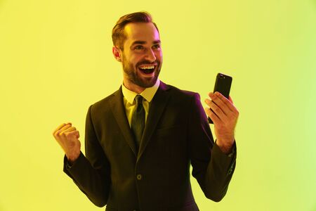 Handsome happy young businessman wearing formal suit standing isolated over yellow background,...