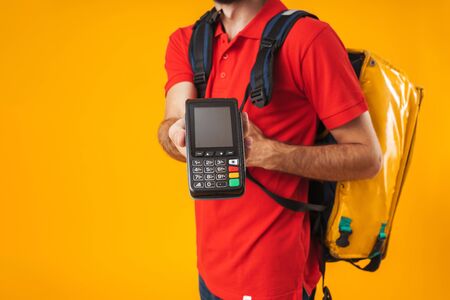Cropped image of caucasian delivery man in red uniform holding payment terminal while carrying backpack with takeaway food isolated over yellow backgroundの写真素材