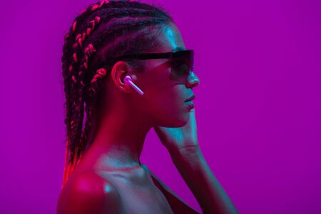 Close up beauty fashion portrait of an attractive young topless african woman wearing dreadlocks standing isolated over purple background, listening to music with earphonesの写真素材