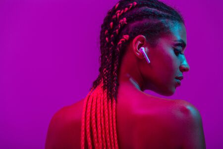 Close up beauty fashion portrait of an attractive young topless african woman wearing dreadlocks standing isolated over purple background, listening to music with earphonesの写真素材