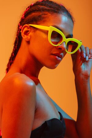 Picture of amazing gorgeous beautiful young african naked woman posing isolated over yellow neon lights wall background wearing sunglasses.の写真素材
