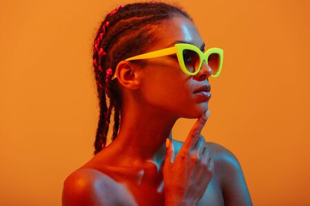 Image of pretty beautiful young african naked woman posing isolated over yellow neon lights wall background wearing sunglasses.の写真素材