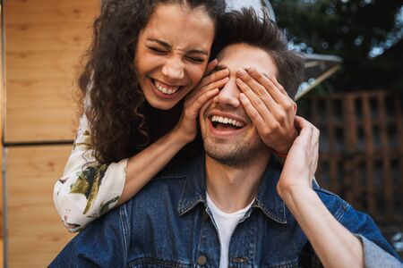 Portrait of brunette laughing woman with curly long hair covering her boyfriend's eyes near trailer outdoorsの写真素材