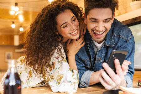 Portrait of happy couple man and woman laughing while using smartphone in cozy kitchen at trailer indoorsの写真素材