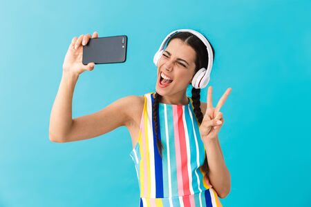 Image of brunette beautiful woman wearing headphones winking while using cellphone isolated over blue wallの写真素材