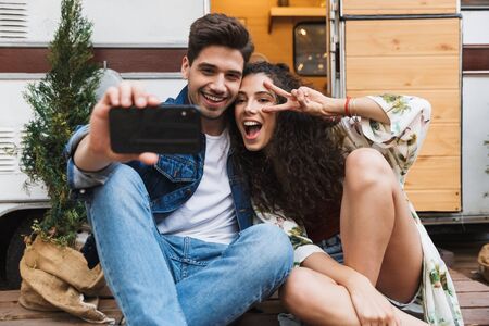 Portrait of joyful couple man and woman taking selfie on cellphone while gesturing peace sign near...