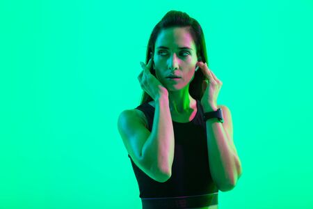 Beautiful young fitness girl standing isolated over green neon background, wearing wireless earphonesの写真素材