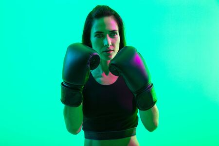 Beautiful confident young fitness girl standing isolated over green neon background, boxingの写真素材