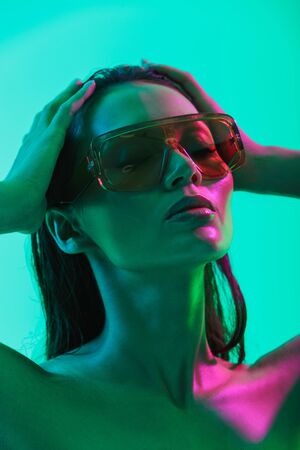 Beauty portrait of an attractive topless young asian woman with long wet hair standing isolated over blue neon background, posing in sunglassesの写真素材