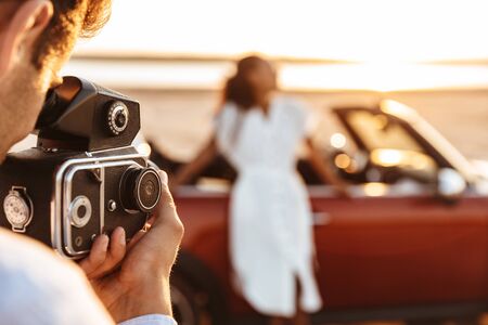 Image of handsome man taking photo on retro camera while woman standing by car at seasideの写真素材