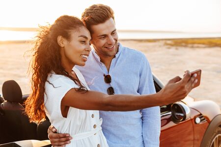 Photo of beautiful multiethnic couple man and woman taking selfie photo on smartphone while standing...