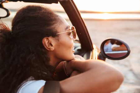 Image of young thoughtful woman smiling and looking aside while riding in convertible stylish car by seasideの写真素材