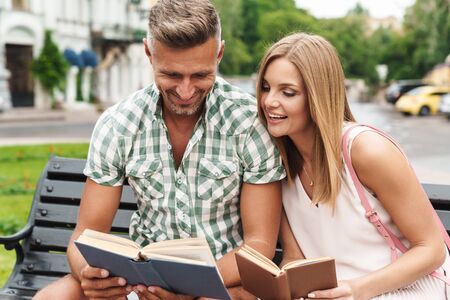 Portrait of beautiful young couple in summer clothes smiling and reading books together while sitting on bench in city streetの写真素材